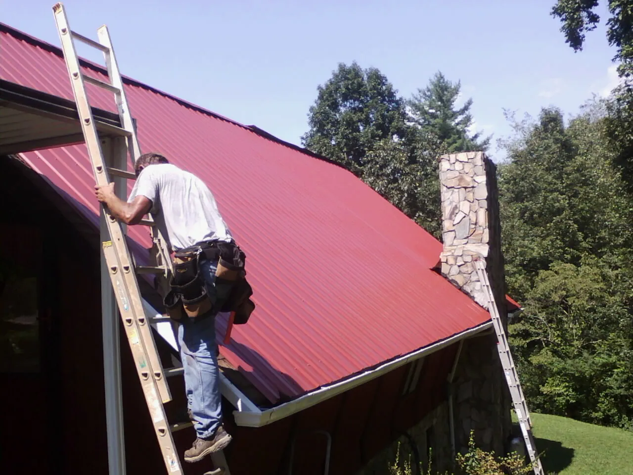 Roofer on a ladder beside a red metal roof and stone chimney — Soffit & Fascia Repair by Roofing Brothers in Grassflat